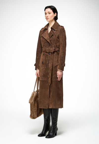 Vrouw draagt lange bruine suède trenchcoat met ceintuur, houdt een pluizige bruine handtas vast en draagt zwarte laarzen tot aan de knie, staand tegen een witte achtergrond.
