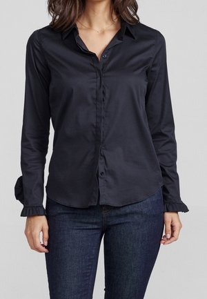 Vrouw draagt een aansluitend zwart blouse met lange mouwen en geruite manchetten en donkerblauwe spijkerbroek, staand tegen een effen grijze achtergrond.