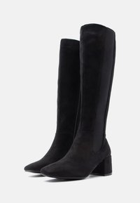 Cizme negre până la genunchi din suede moale, cu vârf ascuțit, toc pătrat și panouri elastice pe laterale pentru a fi ușor de purtat. Închidere cu fermoar pe o parte.