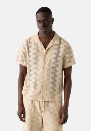 Mann trägt beige kurzärmliges Hemd mit Knöpfen und passende Shorts mit Zickzack-Perforationsmuster, steht vor weißem Hintergrund.