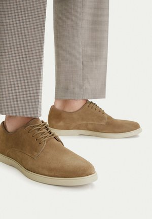 Braune Wildleder-Schnürschuhe, getragen mit hell karierten Anzughosen, auf weißem Hintergrund.