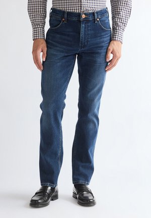 Homme portant un jean droit bleu foncé, des chaussures habillées noires et une chemise à manches longues à carreaux, debout devant un fond uni.