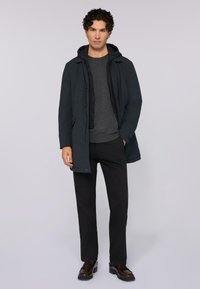 Uomo in piedi con un cappotto blu scuro sopra una felpa nera, un maglione grigio, pantaloni neri e scarpe in pelle marrone scuro, su uno sfondo grigio chiaro.