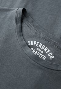 Donkergrijze katoenen t-shirt met ronde hals, met op het binnenlabel in witte letters "SUPERDRY CO., CRAFTED." Soepele textuur.