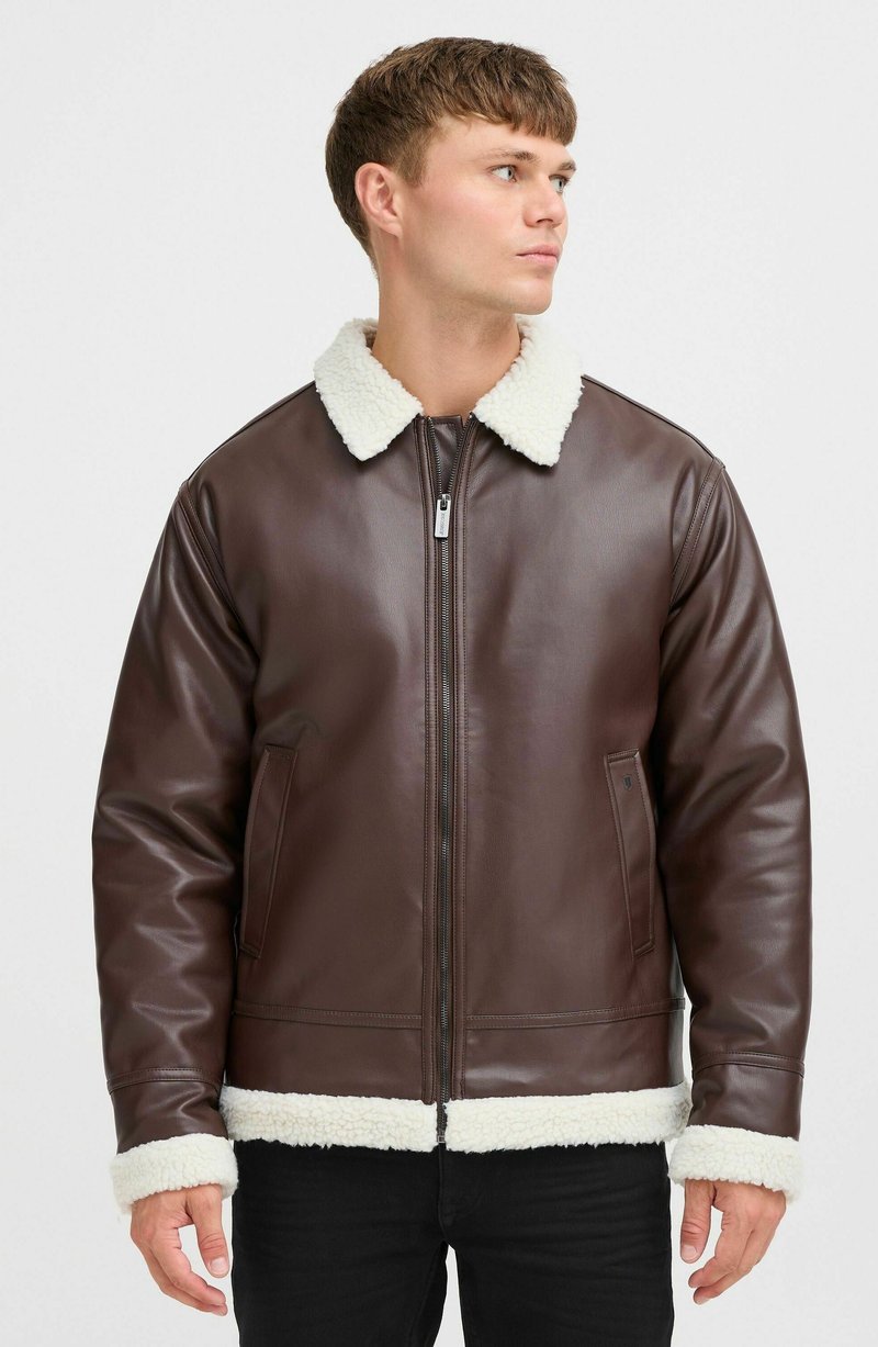 Chaqueta de cuero marrón con un cuello y puños de sherpa blancos, que presenta un cierre frontal de cremallera y dos bolsillos laterales. Textura suave, diseño hasta la cadera.