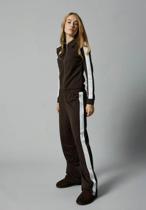 Veste de survêtement zippée marron avec des rayures blanches sur les côtés et un pantalon assorti. Tissu doux avec des poignets et une ceinture côtelés, offrant une coupe décontractée.