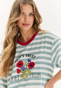Pruhované oversized tričko ve světle modré a bílé, s červeným límečkem, květinovou grafikou a textem "NEVĚŘTE TIŠÍM POLÍM" v černé barvě.