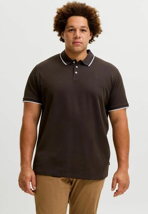 Mann mit lockigem Haar, der ein dunkelbraunes Poloshirt mit weißen Abschlüssen und hellbraune Hose trägt, steht vor einem schlichten Hintergrund.