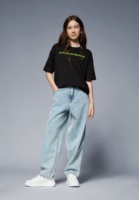 Camiseta de algodón negra con logo en verde neón, combinada con jeans de mezclilla de pierna ancha en azul claro y zapatillas blancas, todo ello sobre un fondo gris.