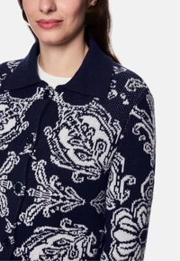 Navy knitted cardigan featuring a white floral pattern, collared neckline, and button closure. Soft texture with a relaxed fit.