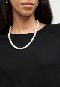 Femme portant un collier simple de perles blanches et un haut noir, montrant le cou et la partie inférieure du visage.