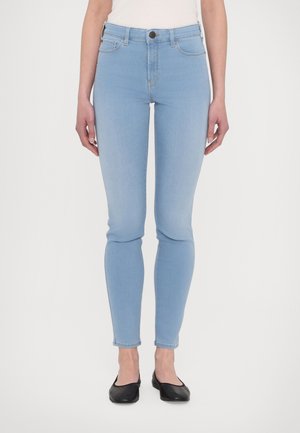 Žena v úzkých svetle modrých džínách, bílém topu a černých balerínkách stojí na hladkém bílém pozadí.