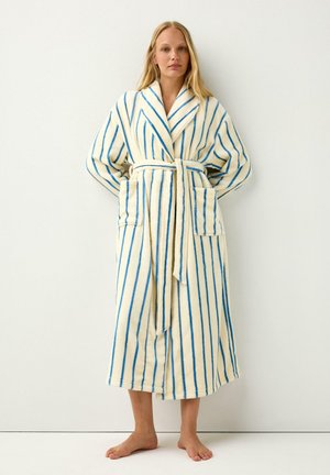 Femme debout pieds nus portant un long peignoir crème avec des rayures bleues verticales, deux poches avant et une ceinture nouée à la taille.