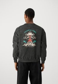 Osoba ubrana w czarną bluzę z kolorowym wulkanicznym wybuchem, palmami i napisem „Tropical Vibes” na plecach.