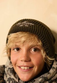 Bonnet en tricot olive avec une texture côtelée, avec un texte brodé blanc. Modèle portant une veste grise en camouflage, souriant.