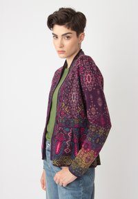 Veste à motifs présentant une base violette profonde avec des designs floraux vibrants en rose, bleu et vert. Silhouette structurée avec des manches longues.