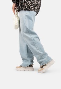 Jean baggy bleu clair, associés à des baskets chunky beiges et noires. Un sac tressé blanc est tenu dans la main gauche. Un haut à imprimé léopard est visible.