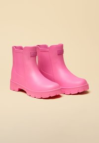 Par korte, knald pink gummistøvler med tyk slidbane og træk-på-stil mod beige baggrund.
