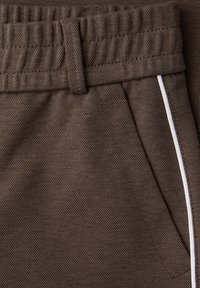 Detailní záběr hnědých texturovaných kalhot s elastickým pasem, poutkem na opasek, boční kapsou a bílým lemováním.