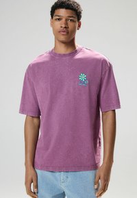 Lila Kurzarm-T-Shirt aus Baumwolle, mit einer kleinen blauen und grünen Blumenstickerei auf der linken Brust. Lässiger Schnitt und Rundhalsdesign.