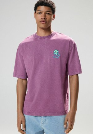 Lila Kurzarm-T-Shirt aus Baumwolle, mit einer kleinen blauen und grünen Blumenstickerei auf der linken Brust. Lässiger Schnitt und Rundhalsdesign.