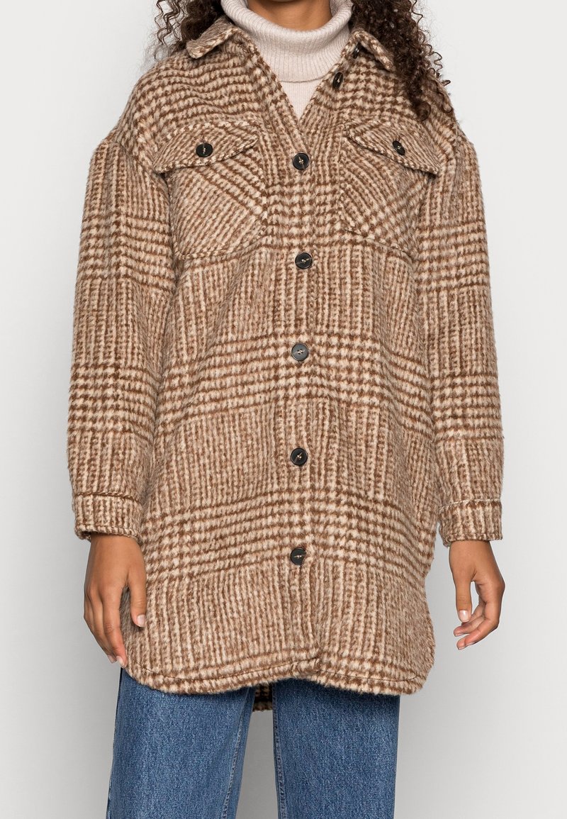 Ruskea ruutukuvioinen overshirt, joka on valmistettu teksturoidusta villasekoitteesta. Siinä on suuret rintataskut, nappisuljenta ja rento leikkaus.