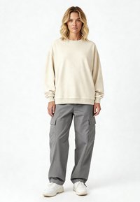 Felpa oversized beige con colletto e polsini a costine, abbinata a pantaloni cargo grigi con tasche laterali, indossata con sneakers bianche.
