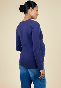 Top de maternidad de manga larga en algodón morado profundo, con un escote redondeado y un diseño ajustado que enfatiza la zona media, combinado con unos jeans.