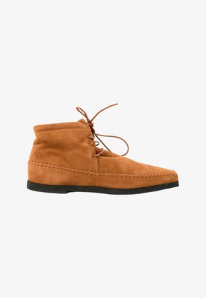 Bottine en daim marron avec lacets à l'avant, détails cousus et semelle plate noire, montrée de profil sur fond blanc.