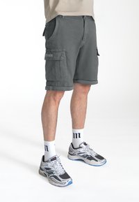 Shorts cargo gris avec ourlet retroussé, poches avant et poches cargo sur les côtés. Portés avec des chaussettes blanches et des chaussures de sport grises. Design simple.