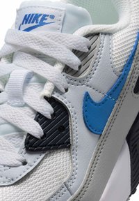 Baskets blanches Nike avec swoosh bleu, lacets blancs et accents texturés noirs, montrant un gros plan du logo sur le côté et la languette.