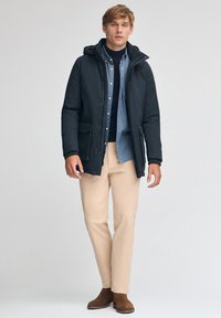 Giacca a vento blu scuro con cappuccio e tasche frontali, indossata sopra una camicia azzurro chiaro, abbinata a pantaloni beige e stivali in suede marroni.