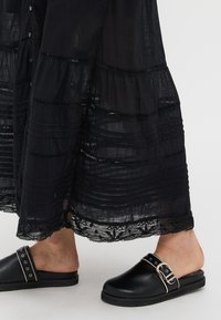 Abito maxi nero a strati con rifiniture in pizzo e dettagli con bottoni, abbinato a zoccoli neri scivolabili con hardware argentato e una cintura con fibbia.
