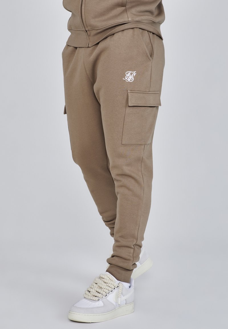 Jogging cargo taupe avec poches à rabat et logo brodé, porté avec des baskets blanches avec des lacets épais style corde.