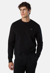 Felpa nera in tessuto liscio con girocollo, polsini a costine e una piccola toppa con logo sul petto. Presenta una vestibilità comoda.