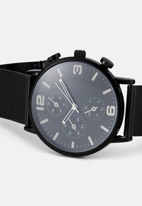 Montre chronographe noire avec un cadran rond gris foncé, index horaires blancs et un bracelet en maille. Elle dispose de trois sous-cadrans et d'un affichage de la date.