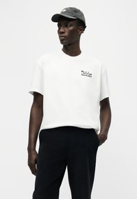 Άνδρας που φοράει λευκό t-shirt Dickies, μαύρο παντελόνι και γκρι καπέλο, στέκεται με το χέρι στην τσέπη, κοιτάζοντας προς τα πλάγια μπροστά σε απλό φόντο.
