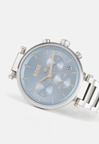 Sølv rustfritt stålklokke med lys blå urskive, tre sub-skiver og aksenter i roségull. Har en datovisning og en metall lenkearmbånd.