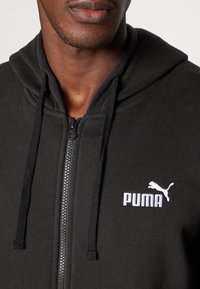 Sort zip-up hoodie lavet af blødt stof, med en kengurulomme, snørehætte og hvidt Puma-logo på brystet.