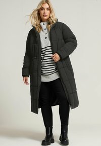 Czarna pikowana długa kurtka z kapturem, noszona na szarym i czarnym swetrze w paski. Model łączy ją z czarnymi spodniami i masywnymi czarnymi butami.
