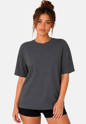 Joven mujer con el pelo castaño recogido en un moño, vistiendo una camiseta lisa de manga corta gris oscura y pantalones cortos negros, de pie frente a un fondo blanco.