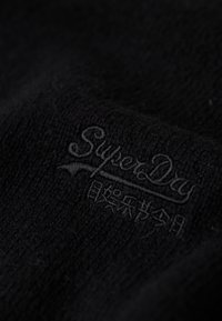 Tessuto a maglia nera con un logo in rilievo "SuperDry" in filo scuro. La texture appare morbida e fine con una leggera lucentezza.