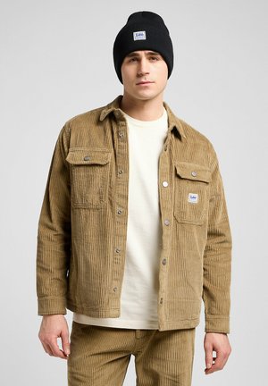 Tan corduroy jacket with vertical stripes, two front pockets, silver buttons, worn over a cream t-shirt, paired with a black beanie.
