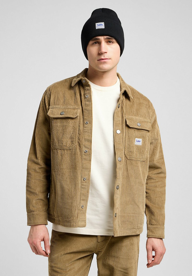 Tanmix corduroy jas met verticale strepen, twee zijzakken, zilveren knopen, gedragen over een crèmekleurig t-shirt, gecombineerd met een zwarte beanie.