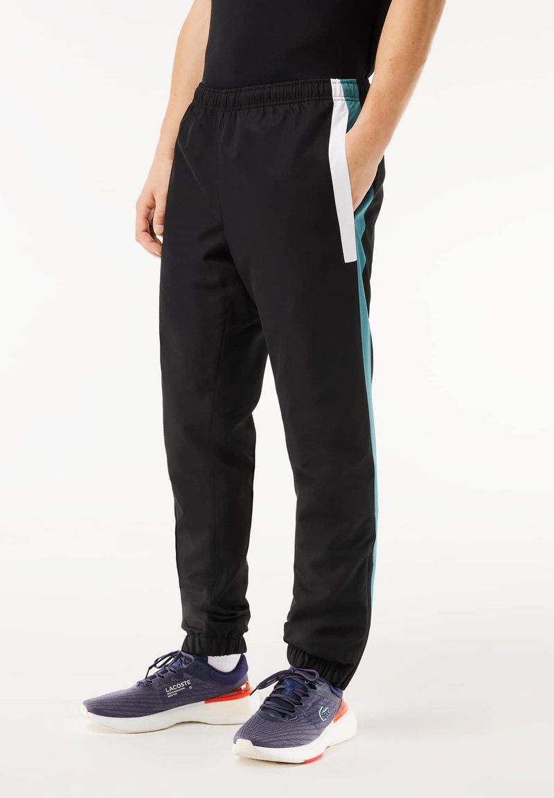 Pantalons de sport noirs avec une taille élastique, rayures latérales en teal et blanc, coupe fuselée et chevilles resserrées, associés à des baskets violettes.