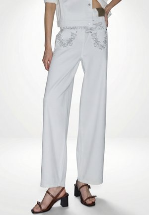 Jean blanc à jambes larges avec poches arrière à motifs, porté avec des sandales marron à talons et une veste blanche courte.