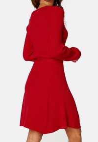 Robe tricotée rouge à côtes, à manches longues, avec un corsage ajusté et une jupe évasée. Elle présente un décolleté rond et un détail de ceinture à la taille.