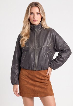 Frau mit langen blonden Haaren, die eine dunkelgraue Lederjacke und einen braunen Wildlederrock trägt, steht vor einem schlichten Hintergrund.
