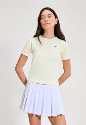 Mladá žena s dlouhými tmavými vlasy, oblečená v světle žlutém krátkém rukávu a bílé plisované sukni, stojící s rukama za zády.