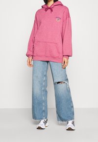 Rosa huvtröja med framficka, med dragsko-stängning och logodetalj. Matchad med slitna ljusblå vida jeans och sneakers.